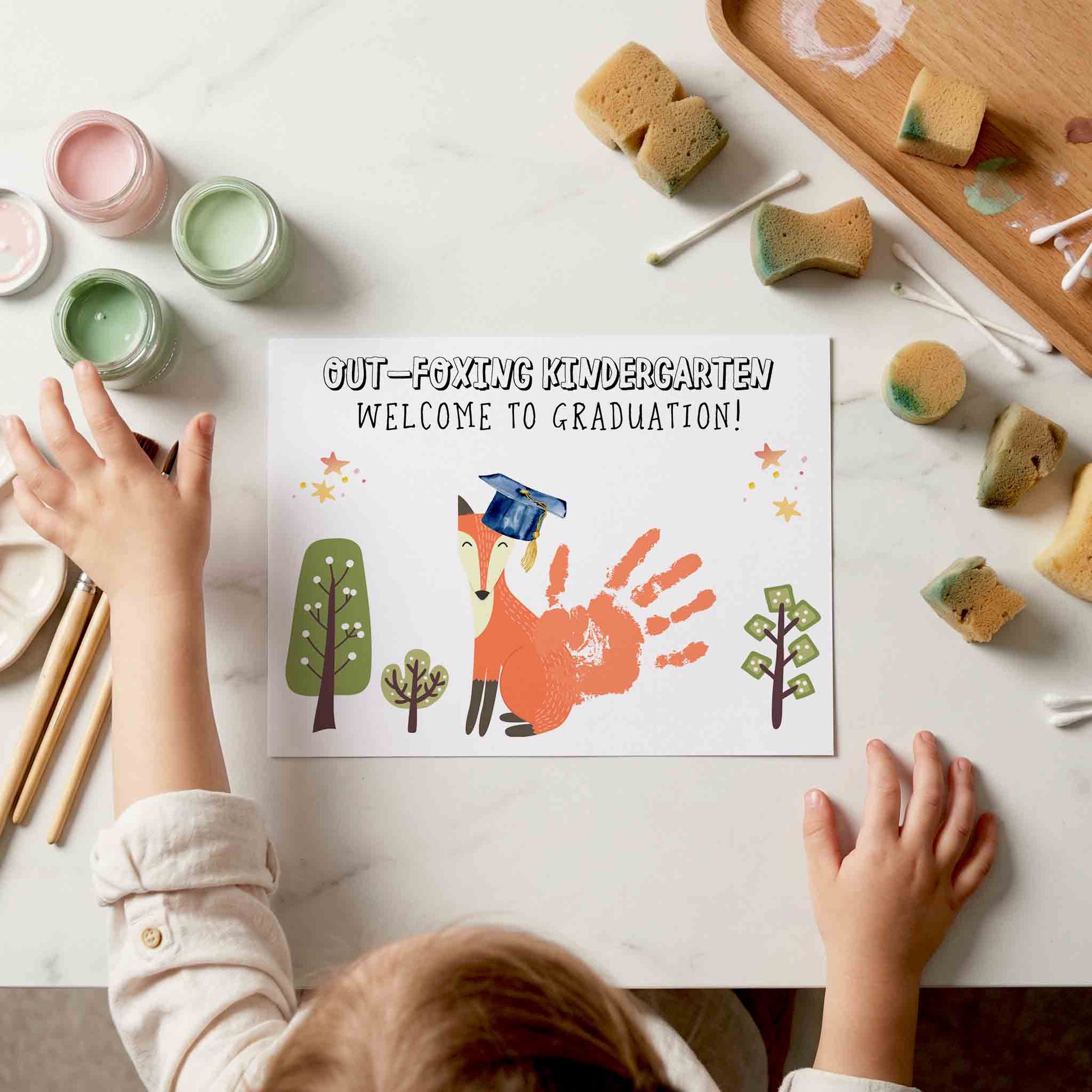 Child's hands reaching towards a graduation card with a fox design on a table with art supplies.