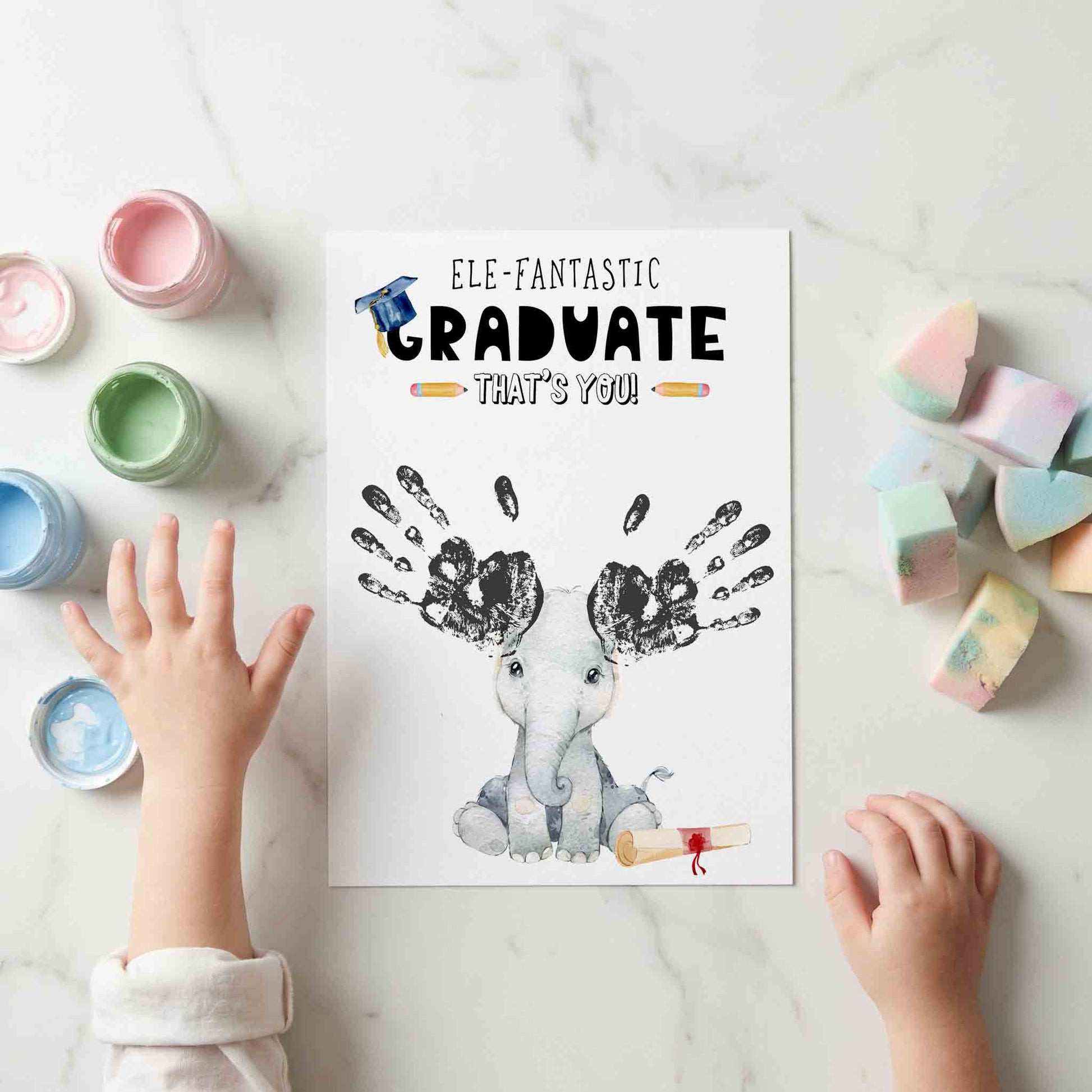 Children's handprint with 'Ele-fantastic Graduate That's You!' card on a marble surface with paint bottles and marshmallows.