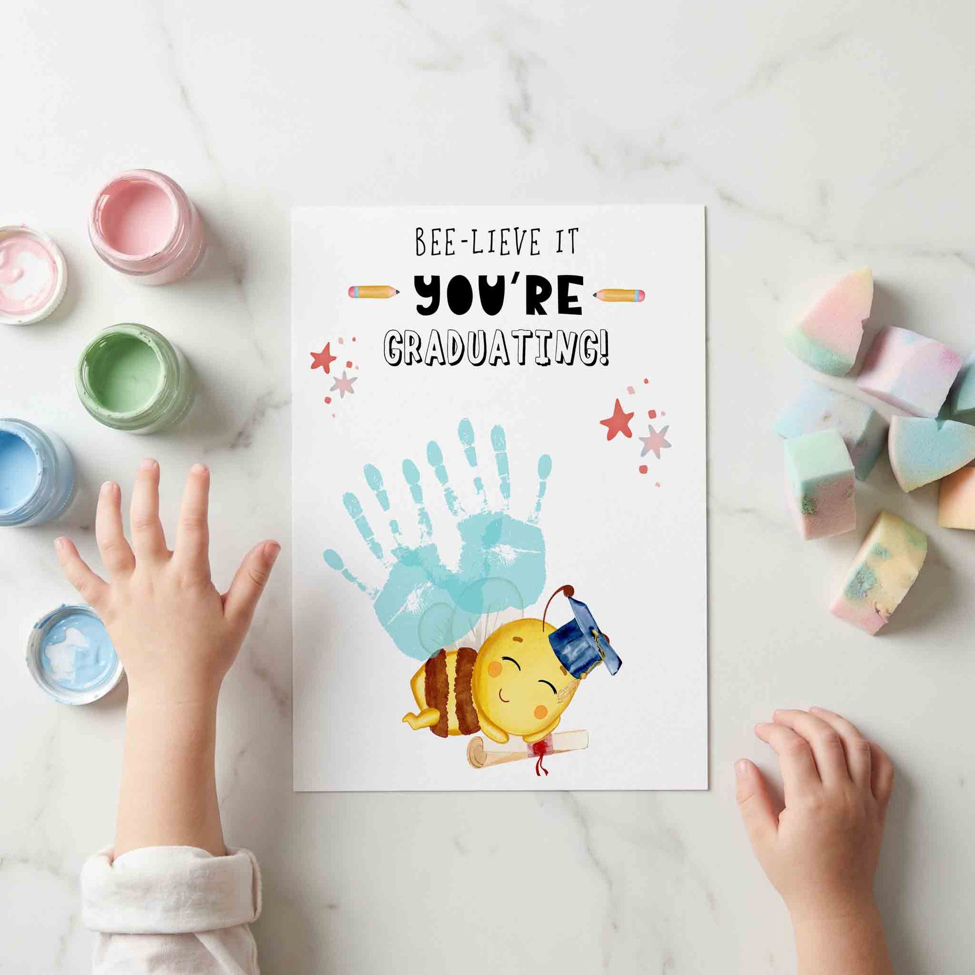Children's hands with handprints on a graduation-themed card on a marble surface.