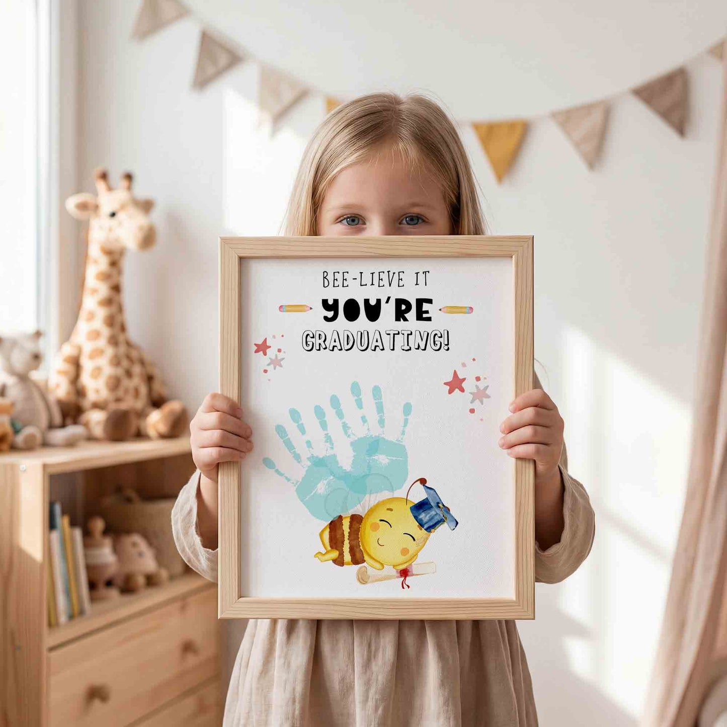 Child holding a framed print with 'BEE-LIEVE IT YOU'RE GRADUATING!' text and handprints.