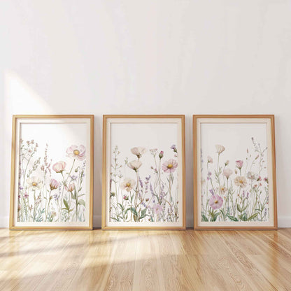 Set of three framed pastel wildflower prints leaning against a wall, featuring pink and lavender flowers on white background, lit by soft daylight.