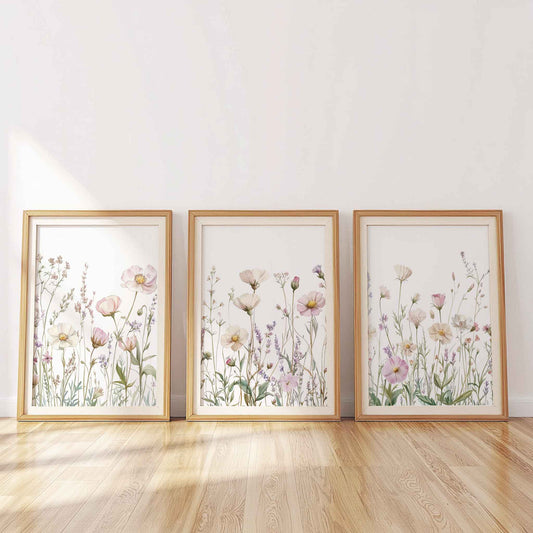 Set of three framed pastel wildflower prints leaning against a wall, featuring pink and lavender flowers on white background, lit by soft daylight.
