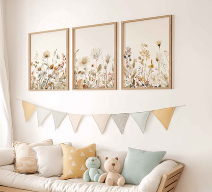 Set of 3 neutral botanical wildflower prints in a baby nursery with wood accents, soft pillows, and pastel flag bunting above a cozy daybed.