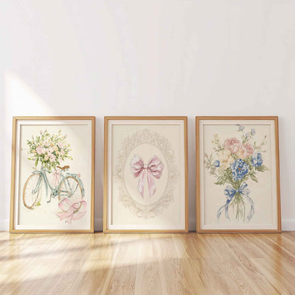 Three framed nursery prints leaning on a wooden floor: a bicycle with pink flowers, a central bow in an ornate frame, and a floral bouquet with blue ribbon.