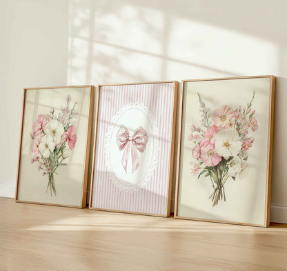 Three framed nursery prints leaning against the wall: two with watercolor pink and white flower bouquets, and one with a vintage pink bow on striped background.